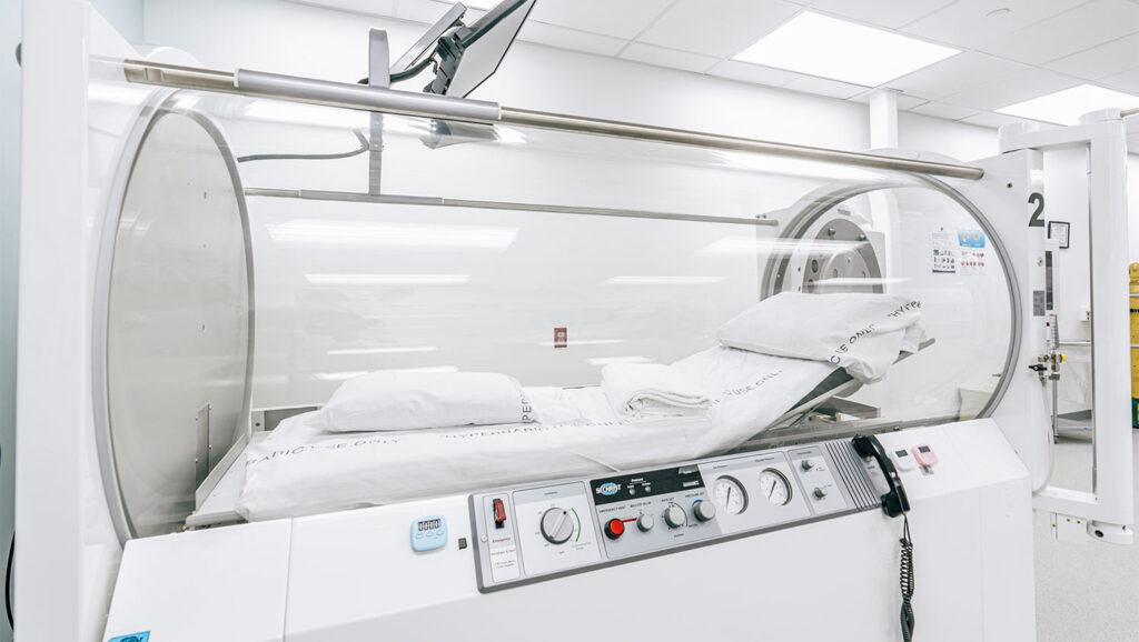 A hyperbaric oxygen therapy chamber with a clear cylindrical enclosure, medical controls, and a bed inside with white sheets and pillows, situated in a clean, modern medical facility.